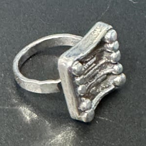 May include: A silver ring with a square-shaped band featuring a textured design and small round accents.