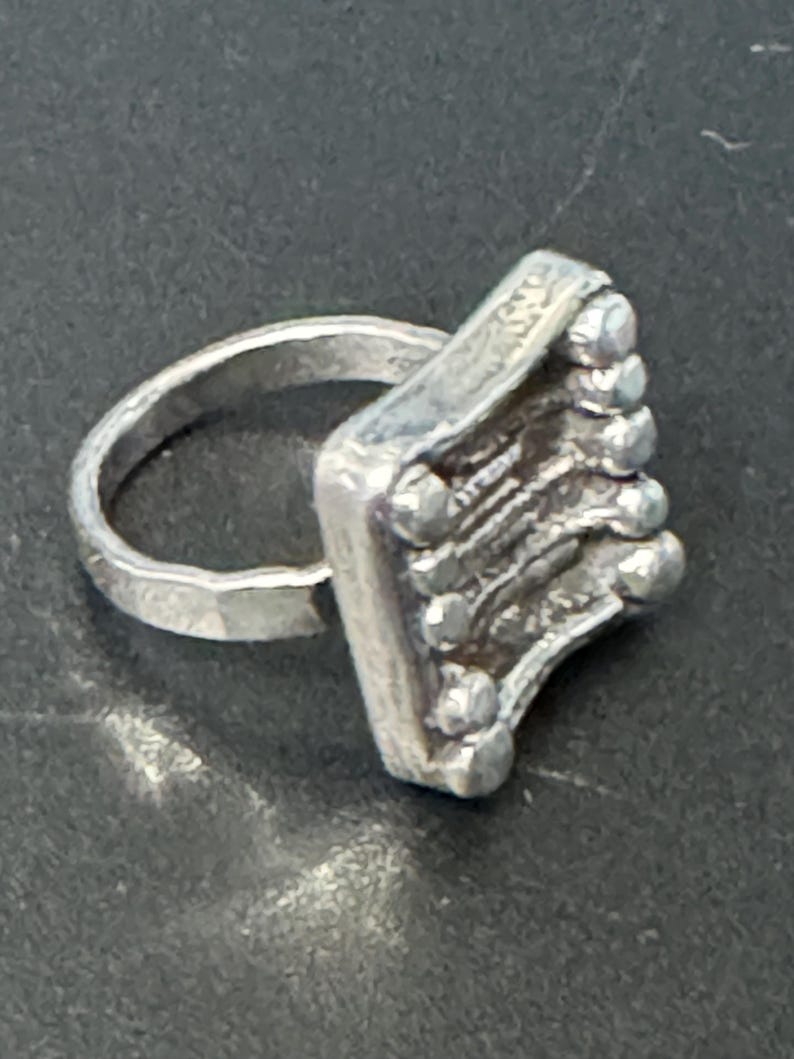 May include: A silver ring with a square-shaped band featuring a textured design and small round accents.