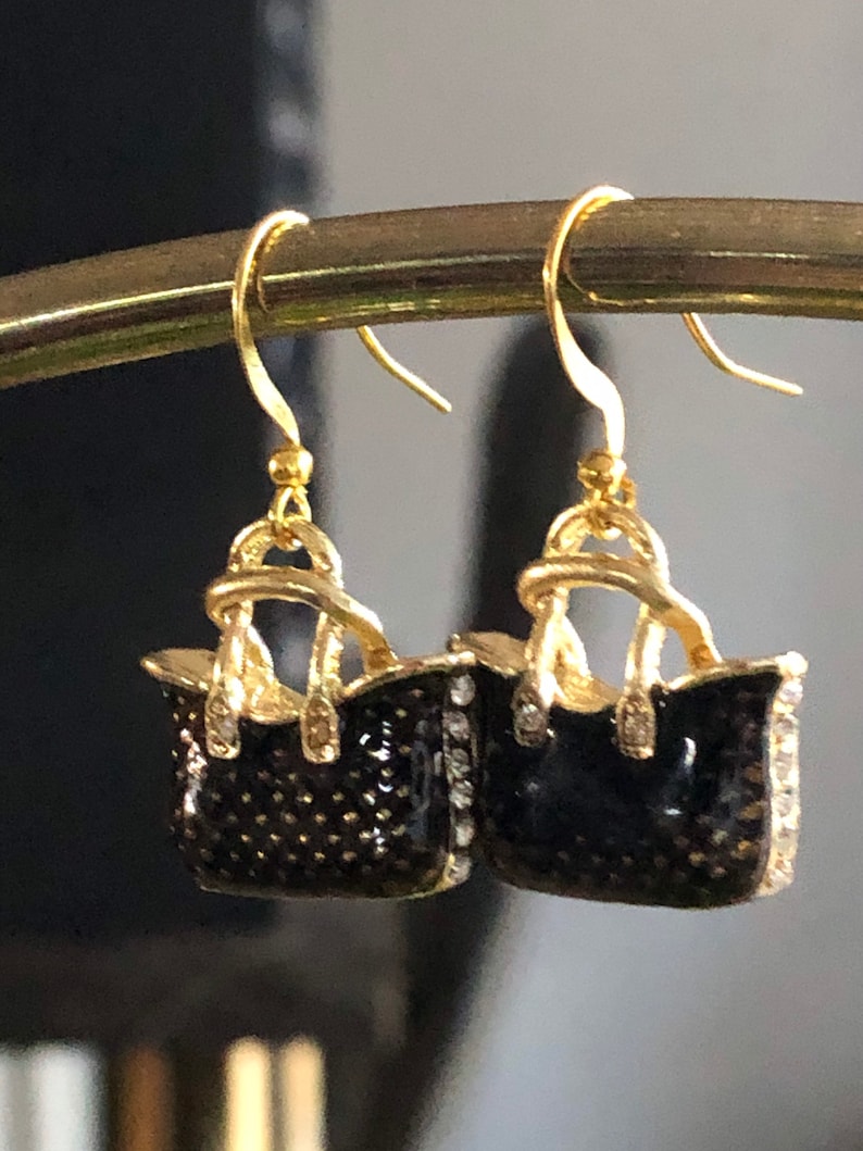 Puede incluir: Pendientes colgantes de tono dorado con un dise&ntilde;o de bolso de mano de esmalte negro y detalles de diamantes de imitaci&oacute;n transparentes.