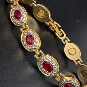May include: A gold-tone link bracelet featuring a series of oval-shaped settings. Each setting contains a vibrant red oval gemstone surrounded by a textured silver-tone border. The bracelet includes a secure clasp mechanism with a circular magnetic closure.