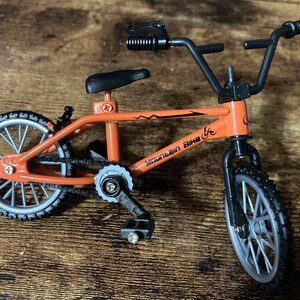 Op de afbeelding: Een kleine oranje mountainbike met zwarte banden en een zwart stuur. De fiets heeft de tekst "Mountain Bike" op het frame. De wielen zijn zilverkleurig met zwarte banden. Het zadel is zwart.