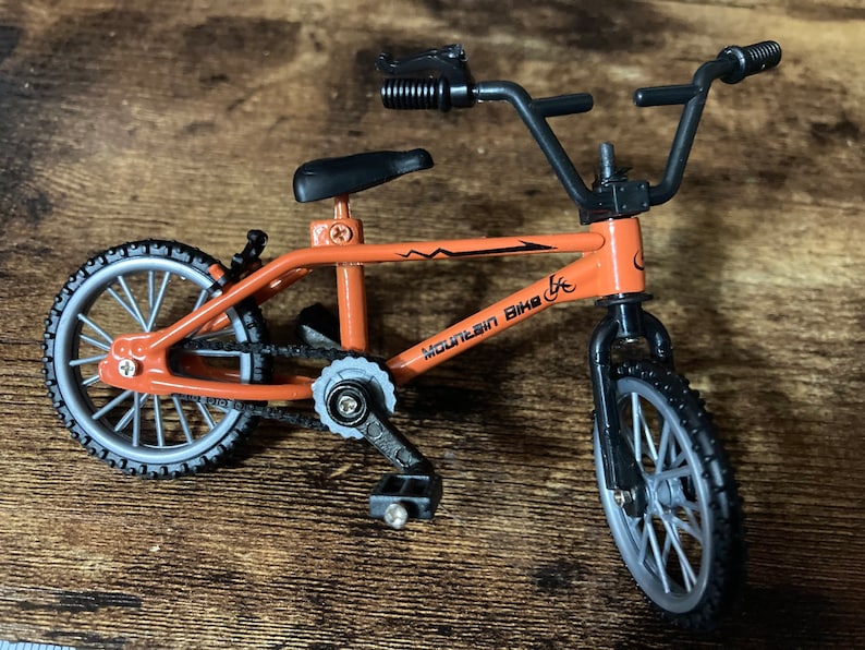 Op de afbeelding: Een kleine oranje mountainbike met zwarte banden en een zwart stuur. De fiets heeft de tekst "Mountain Bike" op het frame. De wielen zijn zilverkleurig met zwarte banden. Het zadel is zwart.