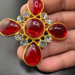 May include: A cross-shaped brooch featuring a central circular red stone surrounded by four teardrop-shaped red stones. The gold-toned metal setting is accented with eight small, clear, round faceted stones placed between the red elements.