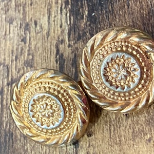 Puede incluir: Un par de pendientes de tono dorado con un diseño circular. Los pendientes tienen un centro elevado y texturizado con un patrón floral.