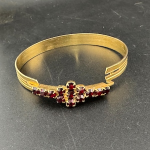 May include: Gold-toned cuff bracelet with a central cluster of red gemstones. The bracelet has a simple, elegant design with a series of parallel lines on the sides. The red gemstones are set in a decorative pattern, adding a touch of color and sparkle.