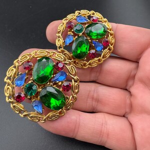 May include: A pair of vintage gold-tone clip-on earrings. Each earring features a circular design with a chain-like border. The center is adorned with green, blue, and red jewel-toned stones. The earrings are set against a dark background.