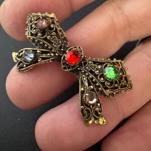 May include: Ornate, antique-style brooch in a bow shape. The gold-toned metalwork features intricate filigree and is adorned with colorful gemstones: red, green, blue, pink, and brown. A vintage-inspired accessory.