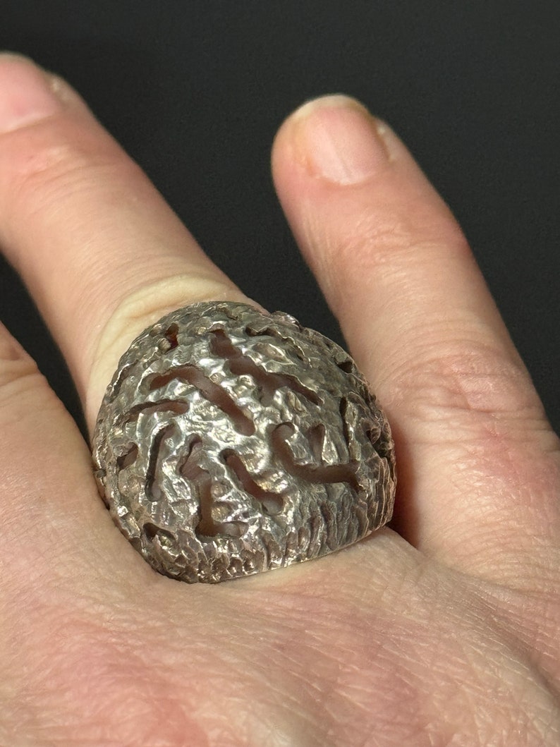 May include: A silver ring with a textured, organic design. The ring is worn on a finger.