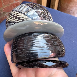 May include: A collection of stacked bracelets in different designs and colours. The top bracelet features a patterned design in black, white, and blue. Below it is a grey bracelet, followed by a carved black bracelet, and a faceted black bracelet.