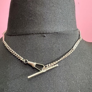 May include: Silver necklace with a T-bar clasp and chain links. The necklace is displayed on a black mannequin head against a pink background. The chain has a delicate, intricate design.
