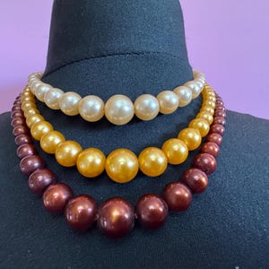 May include: Three-strand necklace featuring graduated faux pearls in cream, gold, and burgundy hues. The necklace is displayed on a black mannequin, showcasing the varying sizes and colors of the beads. A classic jewelry piece.