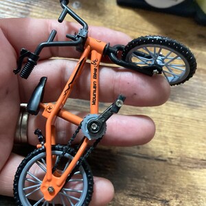 Op de afbeelding: Een oranje miniatuur mountainbike met zwarte accenten. De fiets heeft zwarte banden, een zadel en een stuur. De tekst "MOUNTAIN BIKE" is zichtbaar op het frame. De fiets heeft een ketting en pedalen.