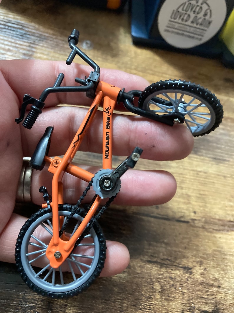 Op de afbeelding: Een oranje miniatuur mountainbike met zwarte accenten. De fiets heeft zwarte banden, een zadel en een stuur. De tekst "MOUNTAIN BIKE" is zichtbaar op het frame. De fiets heeft een ketting en pedalen.
