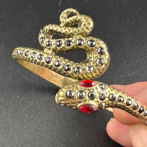 May include: A gold-toned snake-shaped bracelet with black and red gemstones. The snake's head features two red stones, and the body is adorned with small black stones. The bracelet is designed to wrap around the wrist.