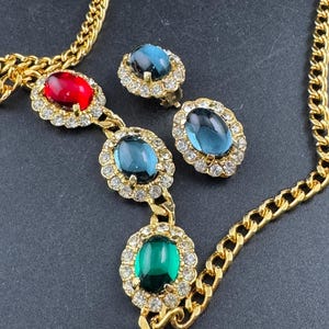 May include: A gold-coloured necklace and matching earrings set. The necklace has a chain with oval gemstones in red, blue, and green, each surrounded by small clear crystals. The earrings feature blue gemstones.