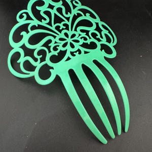 May include: A mint green hair comb with an ornate, openwork design. The comb features a floral motif at the centre and curved, swirling patterns throughout. The comb's teeth are evenly spaced and extend downwards.