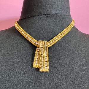 May include: A gold-toned necklace with a decorative tie-like pendant. The necklace features a mesh design with embedded clear crystals. The pendant is composed of three vertical crystal-studded bands. The necklace is displayed on a black mannequin head against a pink background.