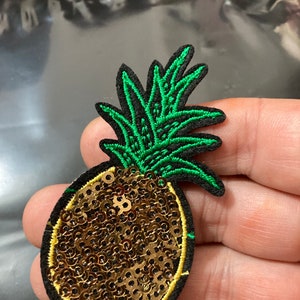 May include: A gold sequin pineapple patch with green embroidered leaves. The patch is perfect for adding a tropical touch to clothing, bags, or other accessories.