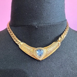 May include: Gold-coloured necklace featuring a triangular blue gemstone pendant. The pendant is framed by small, clear stones. The necklace has a textured, woven chain design, displayed on a black mannequin.