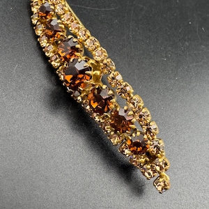 Könnte beinhalten: Eine geschwungene Stab-Brosche mit einer goldfarbenen Metallfassung, verziert mit einer Reihe großer, facettierter bernsteinfarbener Strasssteine, flankiert von zwei parallelen Reihen kleinerer, hellerer champagnerfarbener Kristalle.