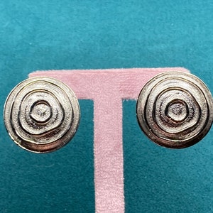 May include: A pair of silver tone earrings with a circular design. The earrings have a concentric circle pattern.