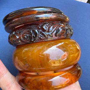 May include: A stack of four amber-colored bangles. The top bangle is a solid, glossy brown. The second bangle features a carved floral design. The bottom two bangles have a marbled appearance. The bangles are held in a hand.