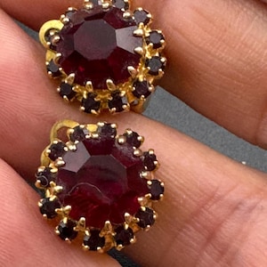 May include: A pair of vintage gold-toned stud earrings. Each earring features a large, faceted, deep red center stone surrounded by smaller, dark red accent stones. The earrings have a classic, ornate design.