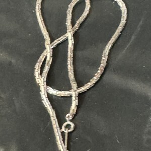 May include: A silver chain necklace with a small, round clasp. The chain is made of small, linked squares.