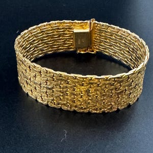 May include: A wide gold bracelet featuring a textured, interlocking woven link pattern. The bracelet has a rectangular box clasp closure that is visible at the top, securing the continuous band of metallic gold links in a uniform design.
