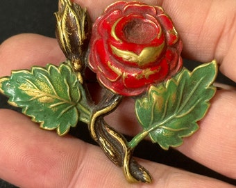 May include: A vintage rose brooch with a red flower, green leaves, and a gold-colored stem. The brooch has a detailed design and is likely made of metal. The rose is in full bloom, with a bud and leaves.