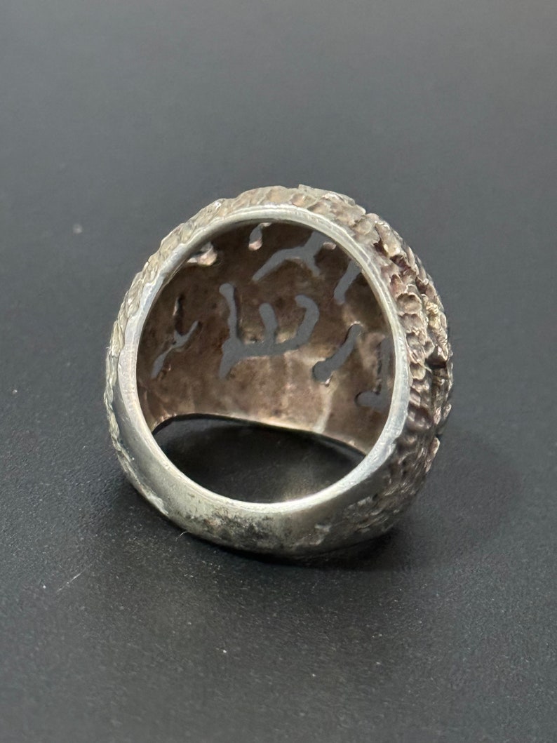 May include: A silver ring with a textured, intricate design. The ring is made of a single piece of metal and has a wide band.