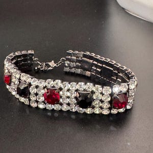 May include: A silver-toned bracelet with a series of square red and black gemstones, interspersed with clear, round crystals. The bracelet is made of multiple rows of small, square links and has a clasp closure.
