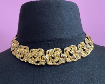 May include: A gold-toned choker necklace with an ornate, textured design. The necklace is displayed on a black mannequin head against a purple background. The necklace features a series of interconnected, irregular shapes.