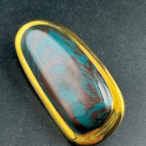 May include: A rectangular brooch with a gold-colored frame. The center features a swirling pattern of teal, brown, and gray colors. The brooch has a polished, glossy finish, and the gold frame adds a touch of elegance.