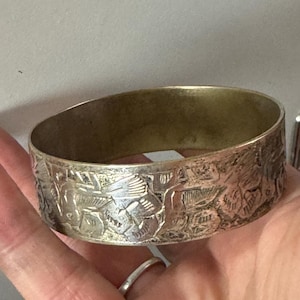 May include: A silver-coloured metal bangle bracelet with an engraved floral design. The bracelet has a wide band and a slightly oval shape. The interior of the bracelet has a gold-coloured finish. The bracelet is held in a hand.