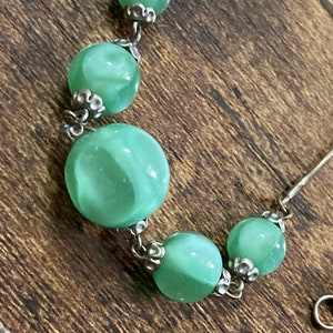 Pu&ograve; includere: Una collana a catena d'argento con cinque perle di vetro verde. Le perle hanno un motivo a spirale e sono collegate da metallo argentato.