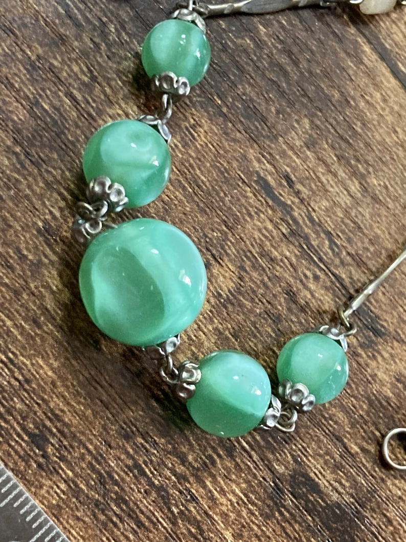Pu&ograve; includere: Una collana a catena d'argento con cinque perle di vetro verde. Le perle hanno un motivo a spirale e sono collegate da metallo argentato.