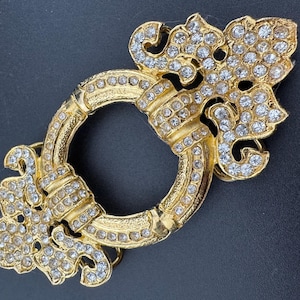 May include: A gold-toned belt buckle with a decorative design featuring a circle surrounded by clear rhinestones. The buckle has a ornate, floral-inspired design.