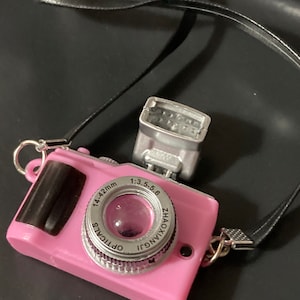 May include: A pink toy camera with a black strap, a silver lens that says "1:3.5-5.6 ZHAOXINGUI OPTICALS 14-42mm", and a silver flash.