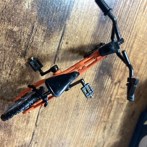 Op de afbeelding: Een miniatuur oranje en zwarte fiets, van bovenaf gezien, met een zwart zadel, stuur en banden. De fiets heeft pedalen en staat op een houten oppervlak. Het frame van de fiets is oranje.