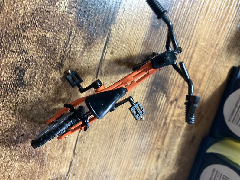 Op de afbeelding: Een miniatuur oranje en zwarte fiets, van bovenaf gezien, met een zwart zadel, stuur en banden. De fiets heeft pedalen en staat op een houten oppervlak. Het frame van de fiets is oranje.