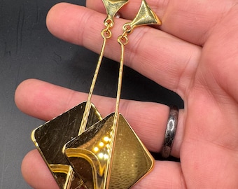 May include: Gold-toned geometric earrings. Each earring features a small triangle at the top, a long slender connecting bar, and a large square with a diamond shape cut out. A small, clear gemstone hangs from the bottom of each earring.