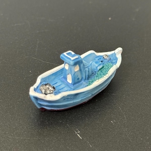 以下が含まれることがあります： 赤色の土台に置かれた、青と白の小さなセラミック製のボートの置物。ボートは白い船室に2つの窓があり、中には緑色の網と白いロープが入っています。