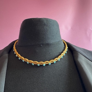 May include: A gold-toned braided choker necklace featuring a series of small, round, light blue stones set along the lower edge. The necklace has a textured, woven appearance and sits against a dark, neutral-colored display form.