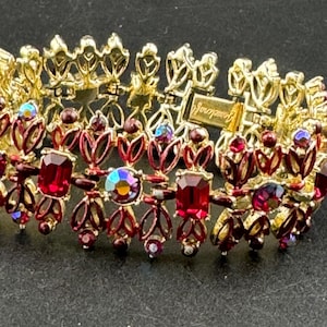 May include: A gold-coloured bracelet with a floral design. The bracelet features red and iridescent gemstones. The clasp is marked "Gemcraft". The bracelet is on a dark surface.