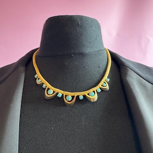 May include: A gold-tone metal collar necklace featuring a textured mesh chain and a series of downward-facing, semi-circular metal drops. Each drop is accented with a small, round, bright turquoise-colored stone set in the center.
