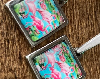 May include: Silver-toned square cufflinks featuring a vibrant flamingo design. The artwork showcases pink flamingos against a turquoise background with floral accents. The cufflinks have a polished finish and a classic, elegant design.