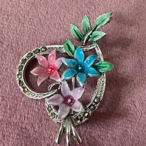 May include: A floral brooch featuring three colorful flowers in pink, blue, and lavender, accented with green leaves. The flowers are set within a silver-toned heart-shaped frame with marcasite detailing. A stylish accessory.
