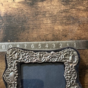 Puede incluir: Un marco de fotos de plata con un dise&ntilde;o ornamentado y un respaldo de terciopelo negro. El marco mide aproximadamente 7,6 cm de ancho y 10,2 cm de alto.
