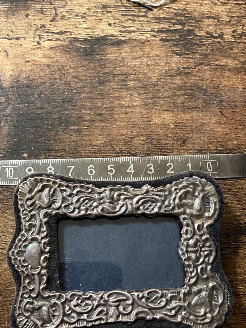 Puede incluir: Un marco de fotos de plata con un dise&ntilde;o ornamentado y un respaldo de terciopelo negro. El marco mide aproximadamente 7,6 cm de ancho y 10,2 cm de alto.